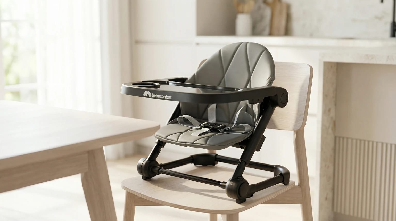 Assento elevatório Bebeconfort Moly em cinzento e preto, colocado numa cadeira de jantar de madeira clara junto a uma mesa, numa cozinha moderna e luminosa.
