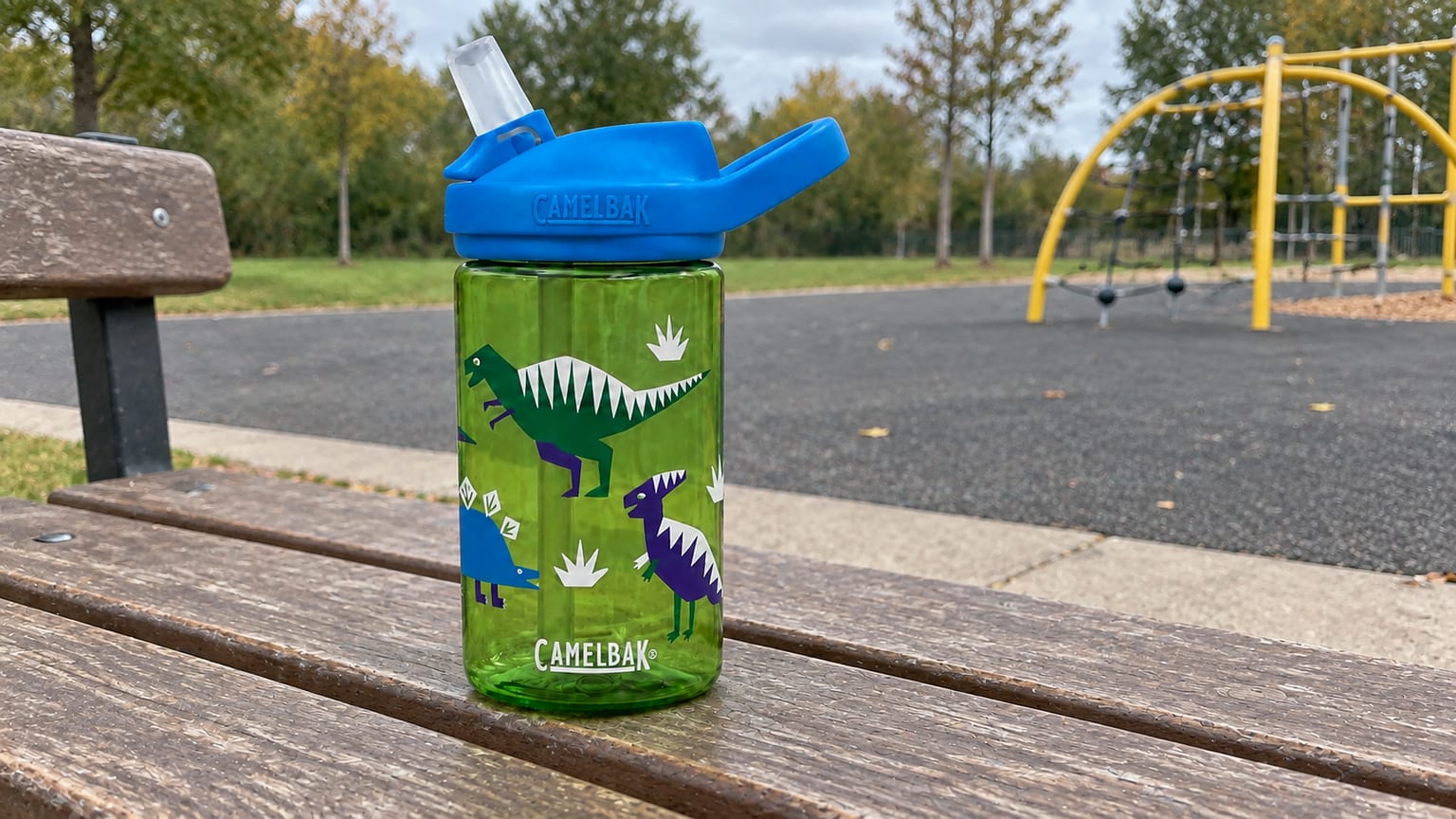 Garrafa CamelBak verde com dinossauros e bocal levantado, num banco junto de um recreio ao ar livre.