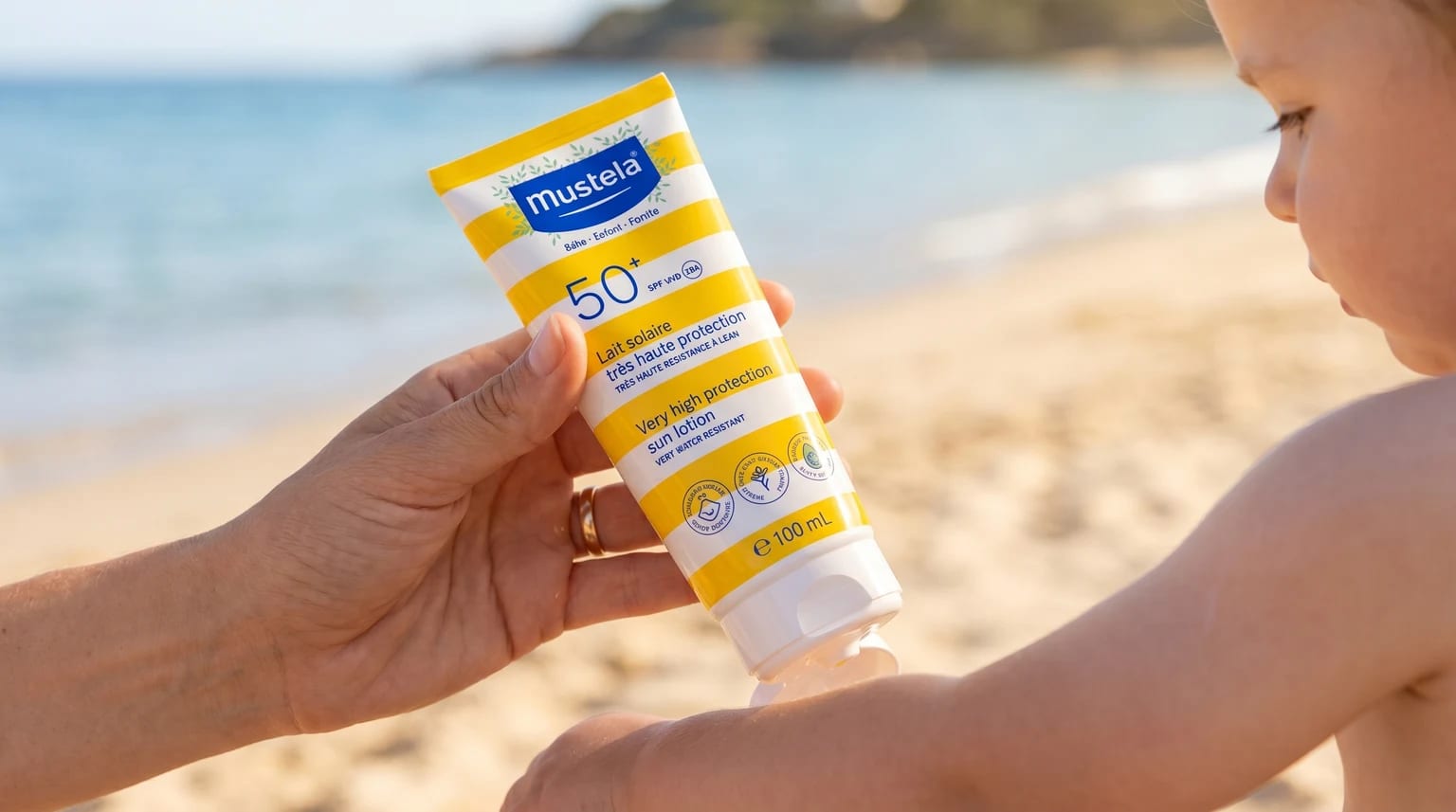 Tubo de protetor solar Mustela SPF 50+ a ser segurado pela mão de um adulto, pronto a ser aplicado na pele de uma criança, com uma praia soalheira e calma como fundo.