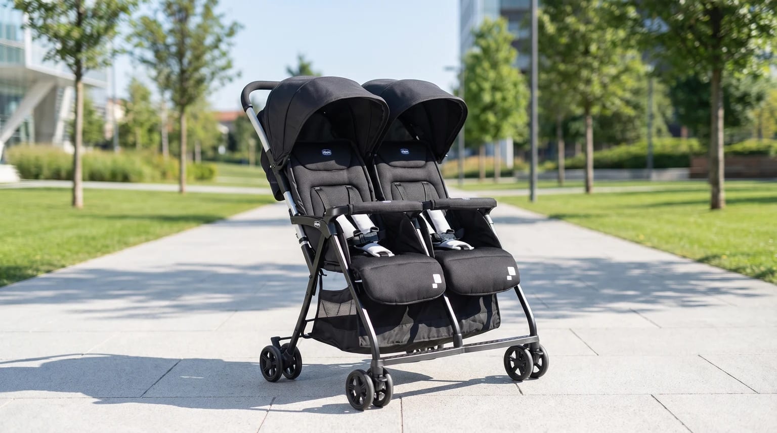 Carrinho de bebé duplo Chicco OHlalà Twin preto, vazio e estacionado num caminho soalheiro de um parque moderno.