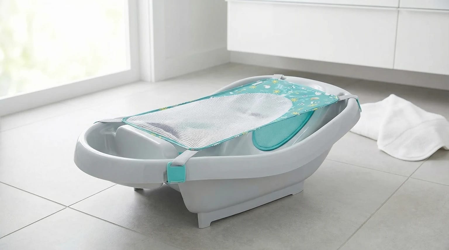 Banheira de bebé Ingenuity cinzenta clara com um suporte de rede turquesa, pousada no chão de uma casa de banho moderna e luminosa.