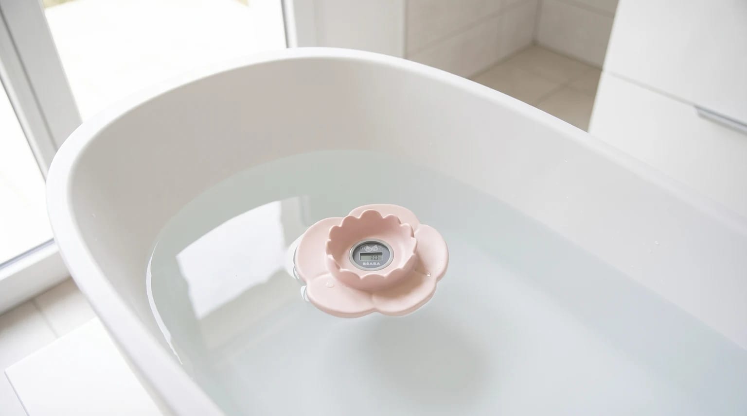 Termómetro digital Béaba Lotus rosa em forma de flor pousado na água calma de uma banheira de bebé branca, numa casa de banho luminosa.
