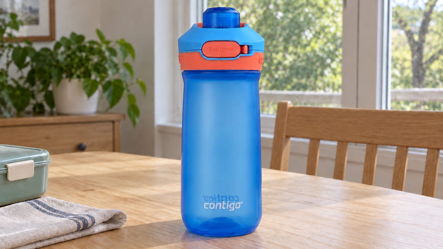Garrafa Contigo azul com tampa coral, numa mesa junto de uma lancheira, com uma janela ao fundo.