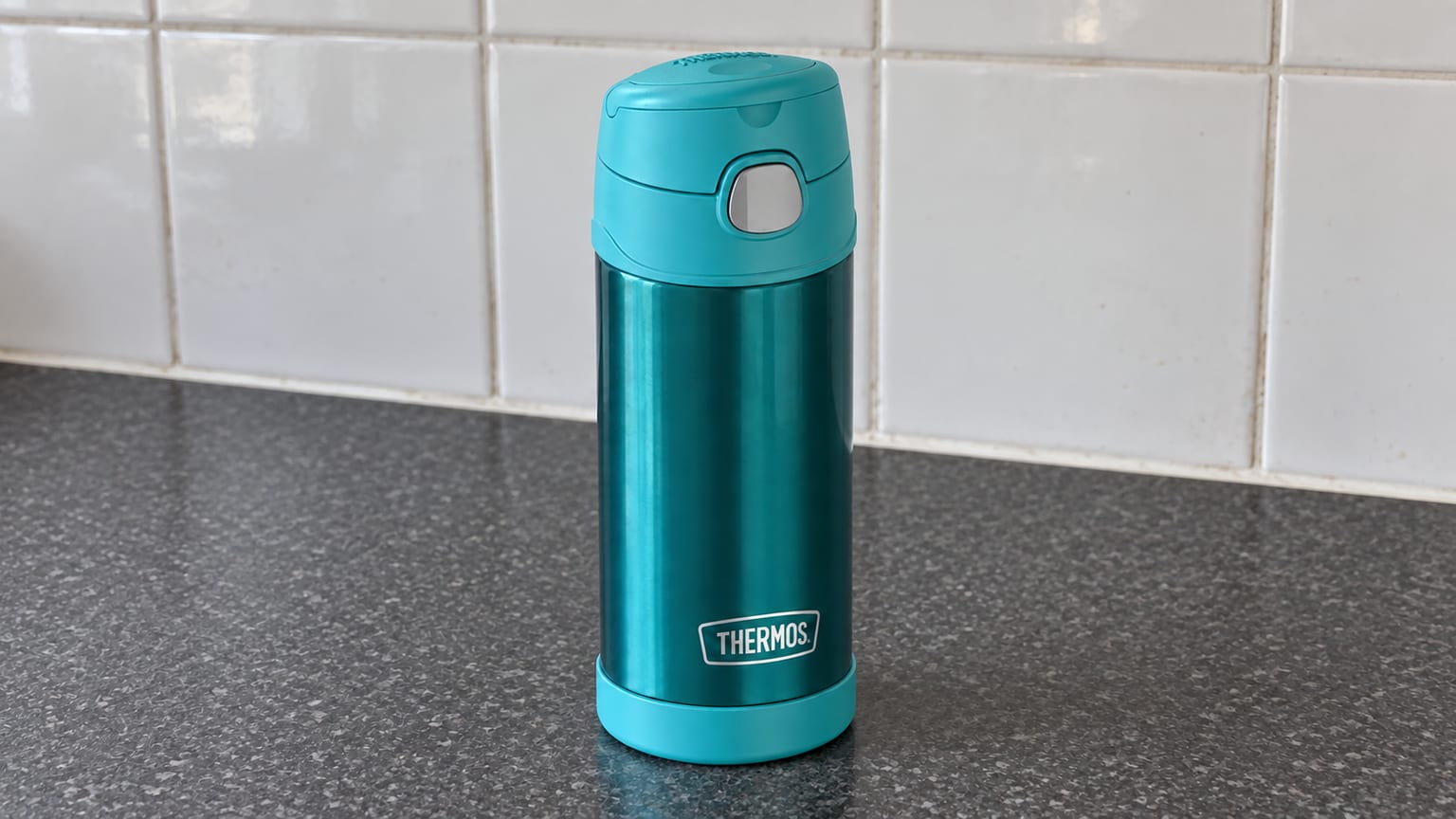 Garrafa Thermos azul-petróleo com tampa fechada, sobre uma bancada cinzenta diante de azulejos brancos.